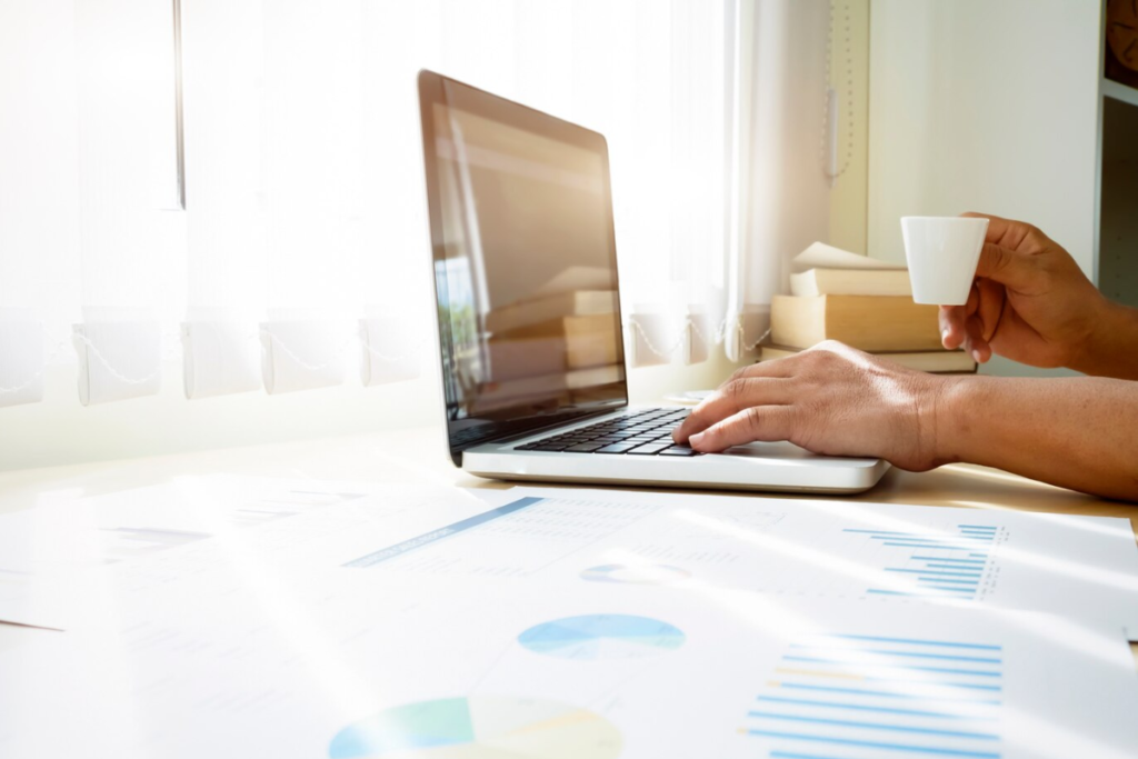 Person holding a cup of coffee while working on a laptop, reviewing printed SEO results with graphs and charts
